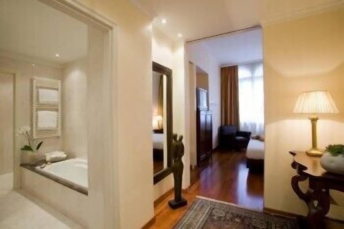 A hotel room with a large bed, a sitting area with a sofa and armchairs, a small table with a lamp, and a view of the hallway.