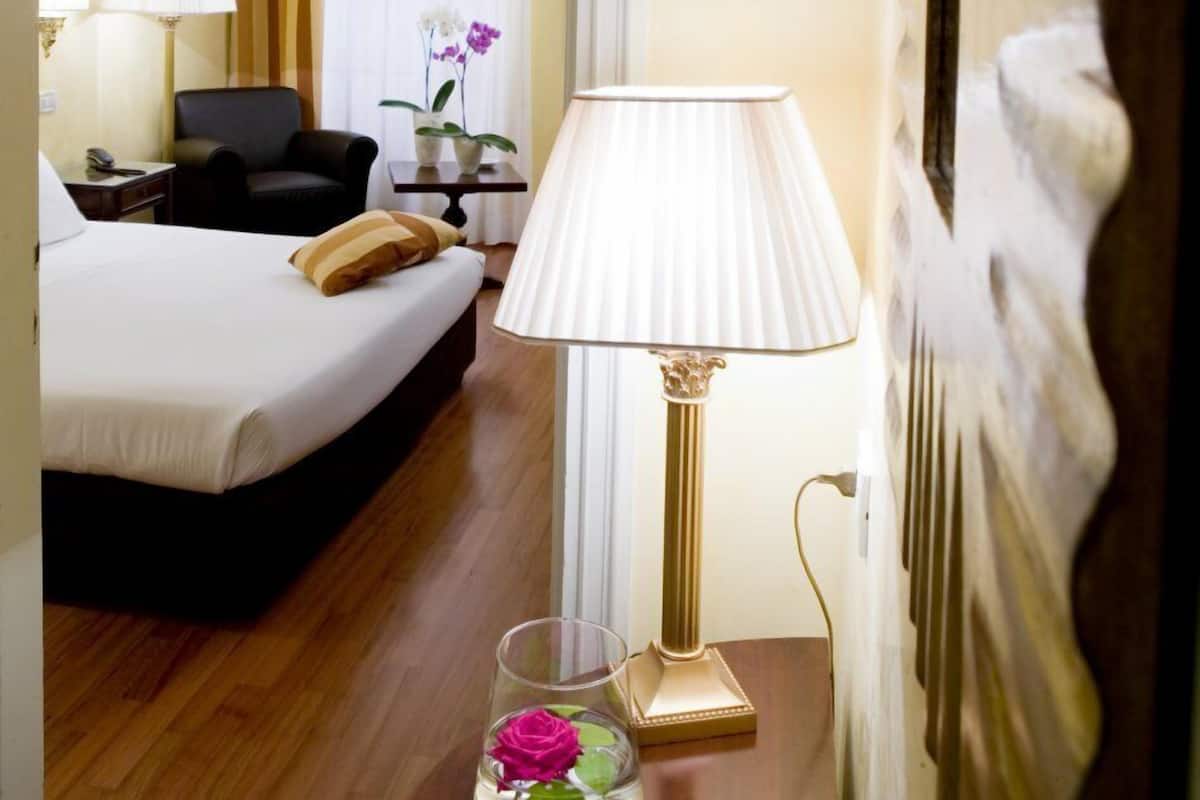 A hotel room with a bed, a chair, a lamp, and a vase with flowers.