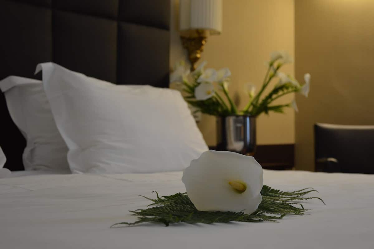 A neatly made bed with white linens, a white flower centerpiece, and a vase with white flowers in the background.