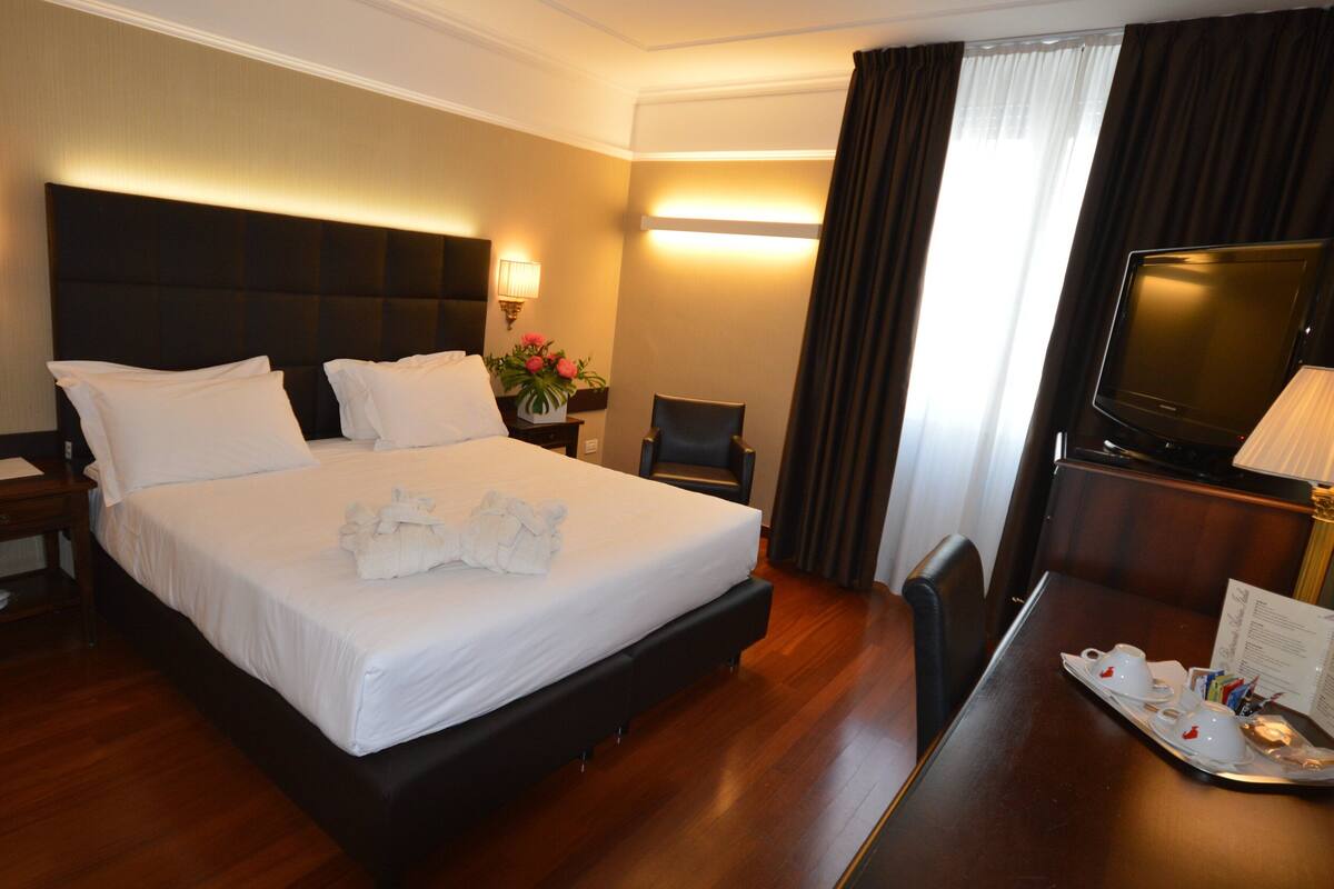 A hotel room with a large bed, a desk with a tray of cups, a chair, a television, and a nightstand with a lamp.