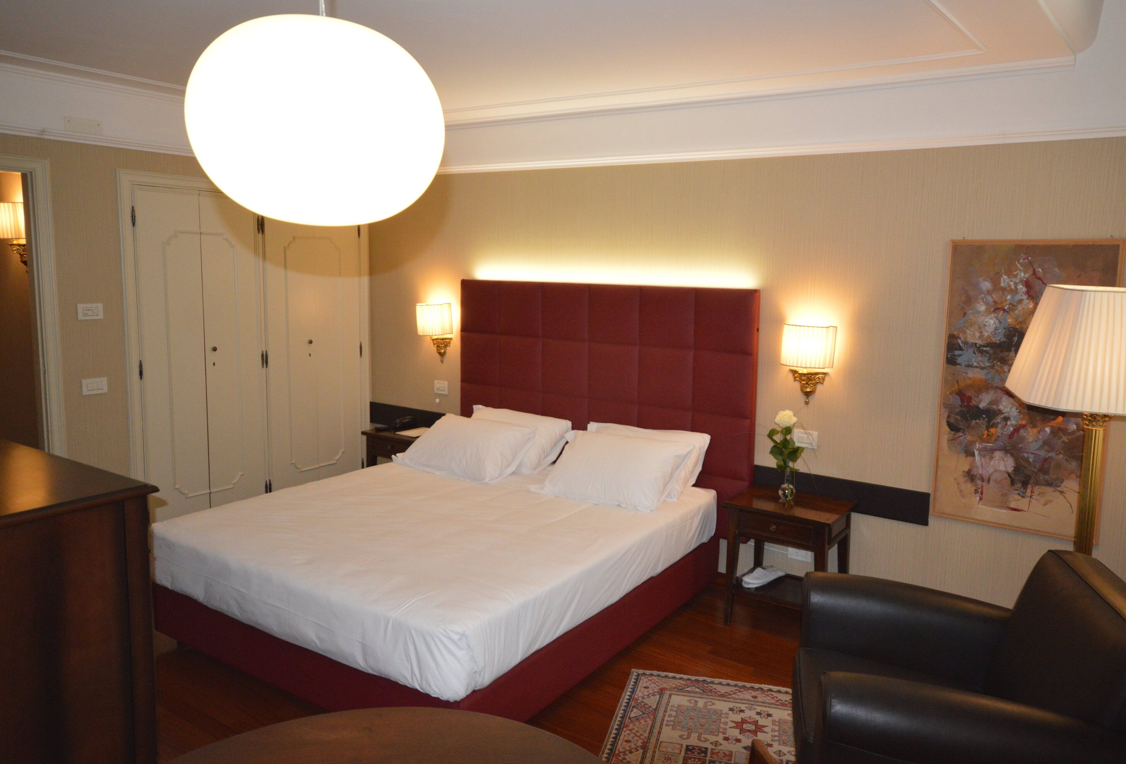 A hotel room with a large bed, a red headboard, two bedside tables with lamps, a wooden dresser, and a painting on the wall.