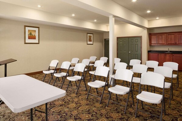 Un salón de conferencias con sillas plegables blancas dispuestas en filas, una mesa blanca y una alfombra estampada.