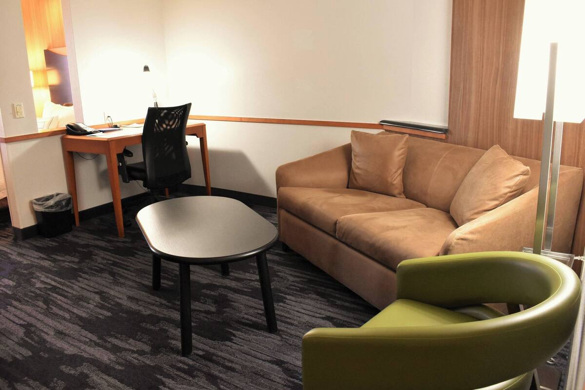 Een hotelkamer met een bruine bank, een groene fauteuil, een kleine tafel, een bureau met stoel en een lamp.