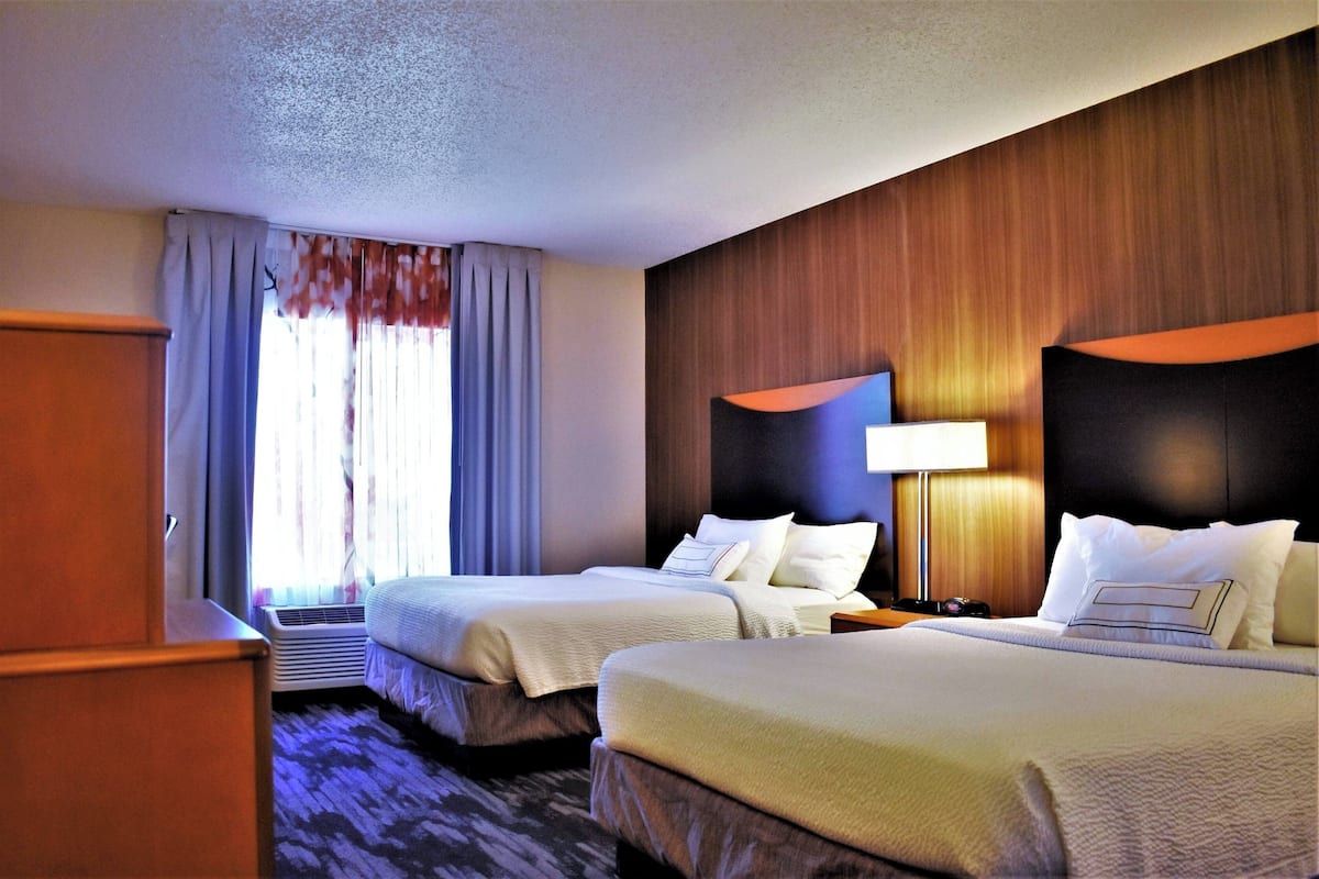 Een hotelkamer met twee bedden, een houten hoofdbord, een nachtkastje met een lamp en een raam met gordijnen.