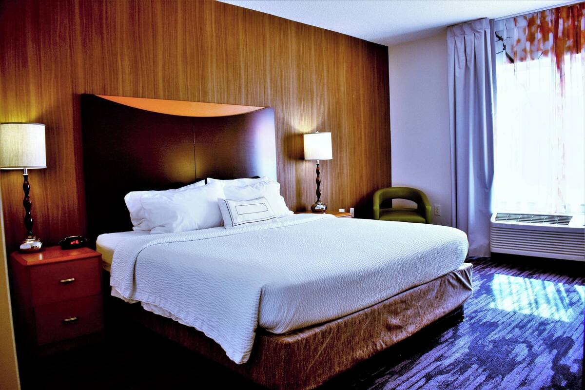 Een hotelkamer met een groot bed, een houten hoofdbord, twee nachtkastjes met lampen, een stoel en een raam met gordijnen.