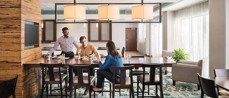 Un comedor moderno de hotel con una larga mesa, múltiples sillas y un amplio ventanal que proporciona luz natural.