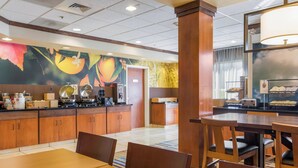 A hotel dining area with wooden tables and chairs, a kitchen counter with appliances, and a mural with fruit designs on the wall. - Fairfield Inn & Suites by Marriott Columbia (Columbia)