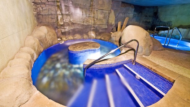 Un jacuzzi con un muro de piedra de fondo, una plataforma de madera y una escalera metálica.