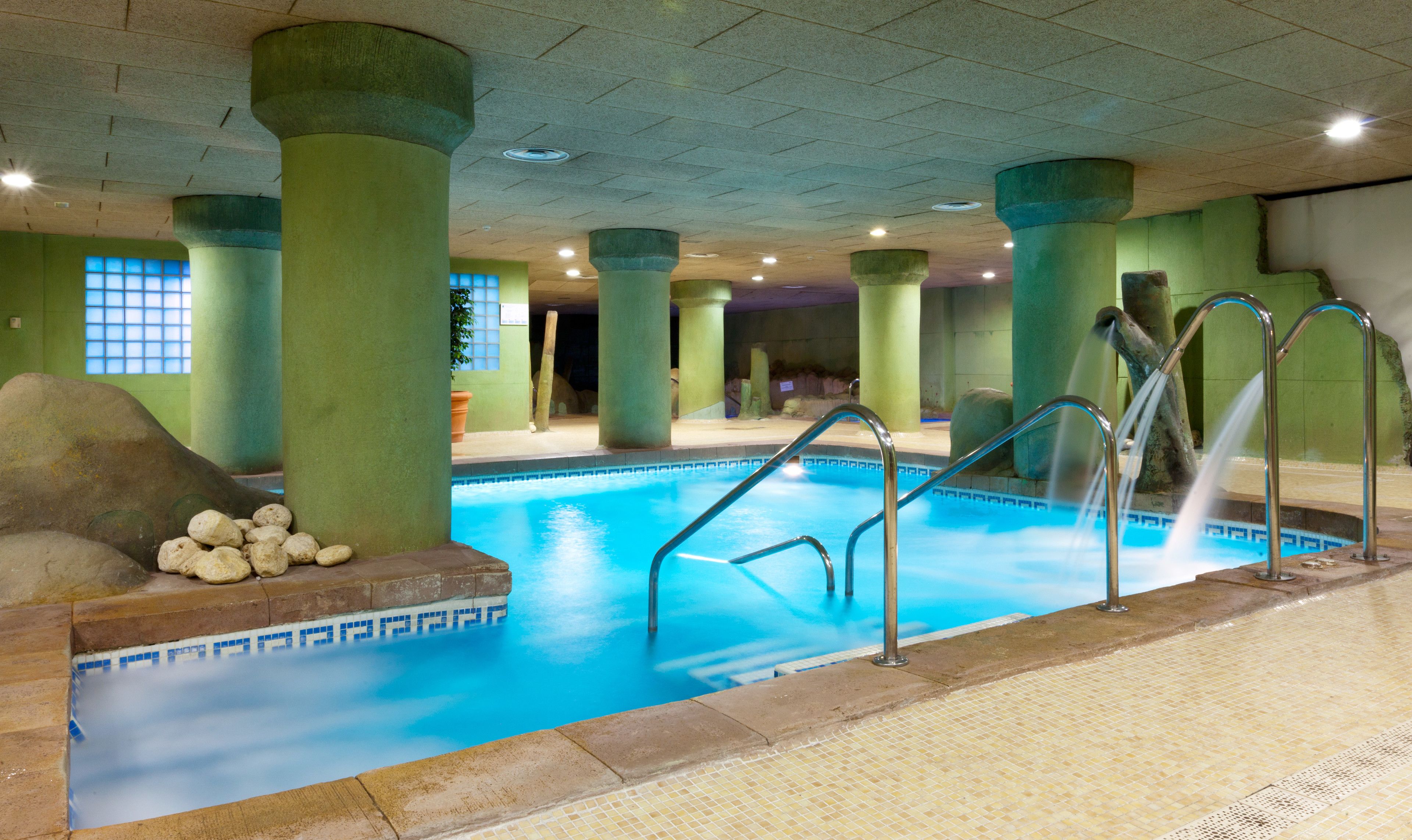 An indoor swimming pool with clear blue water, surrounded by green pillars and a tiled floor.