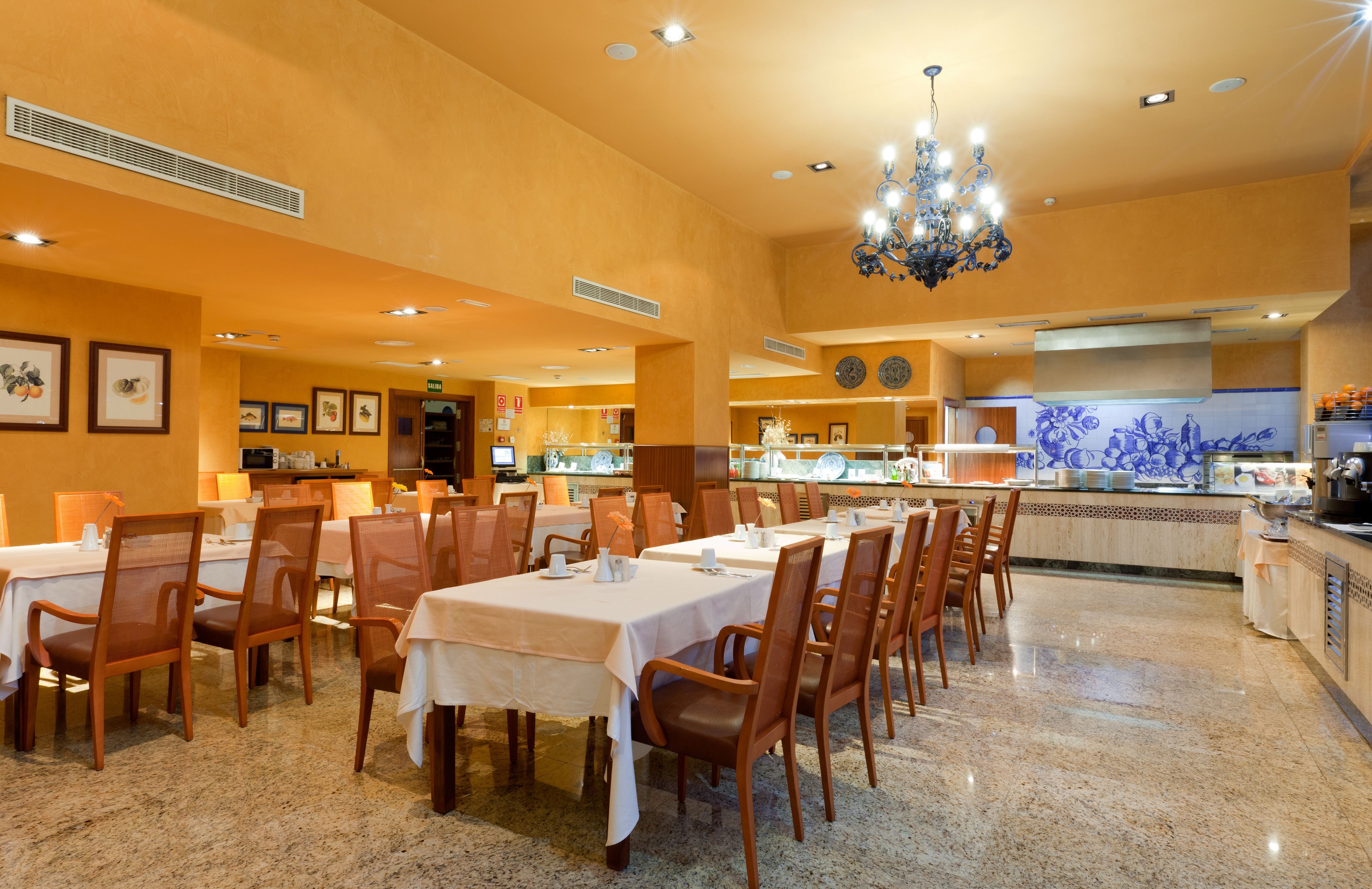 A restaurant with multiple tables and chairs, a chandelier, and a kitchen area in the background.