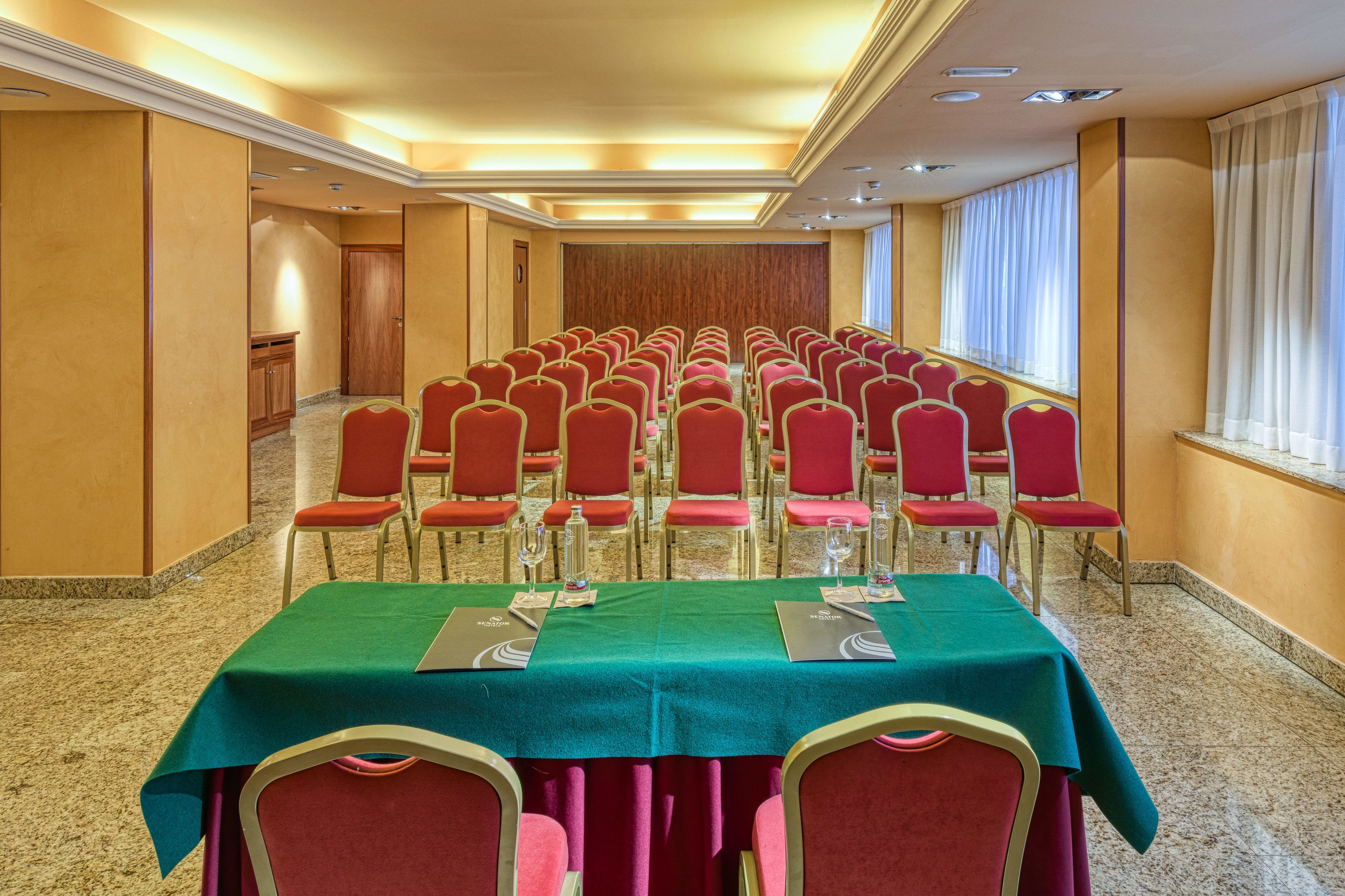 Een congreszaal met rijen rode stoelen, groene tafels en een wand van houten panelen.