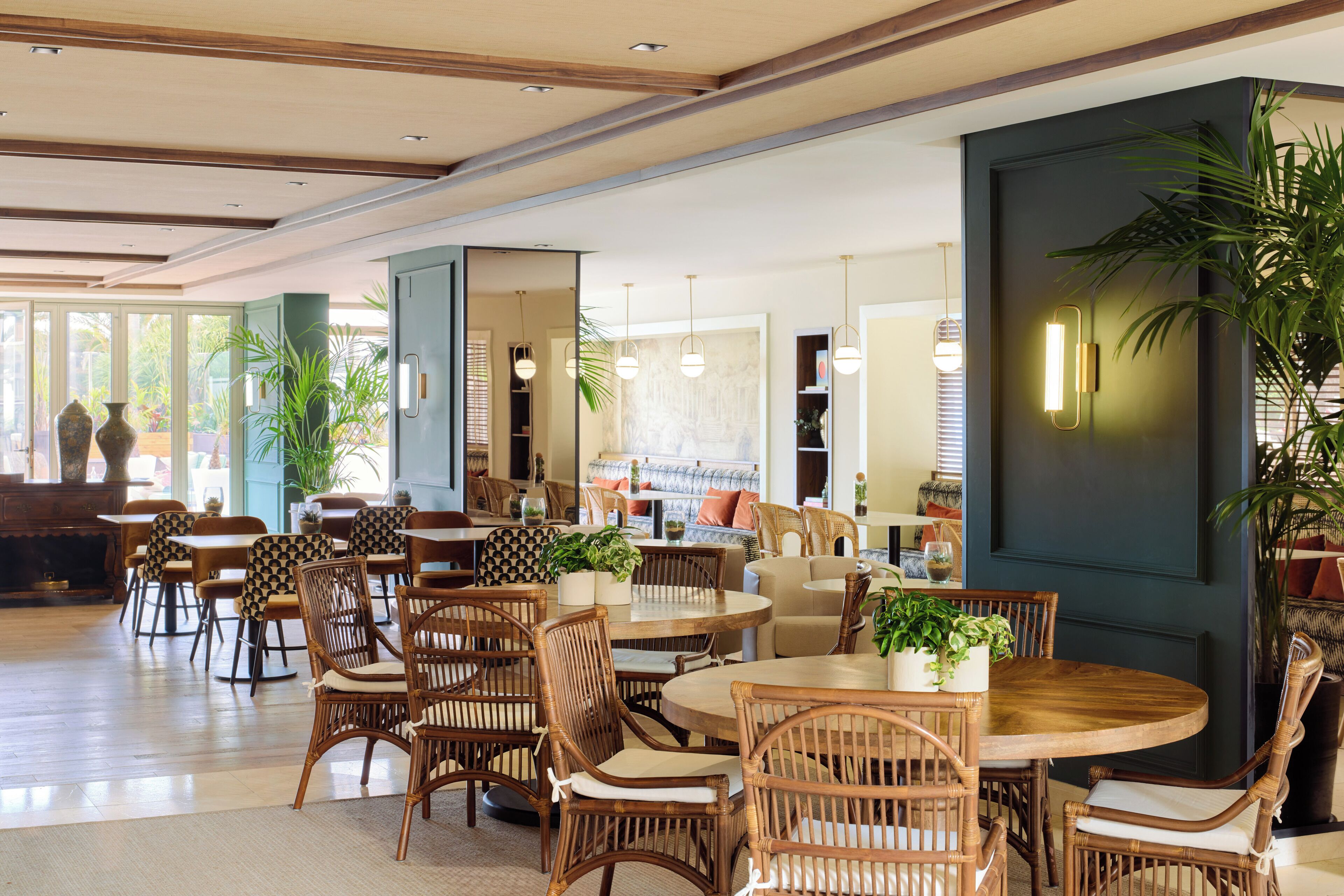 A spacious indoor area with multiple round tables and wicker chairs, surrounded by large windows and indoor plants.