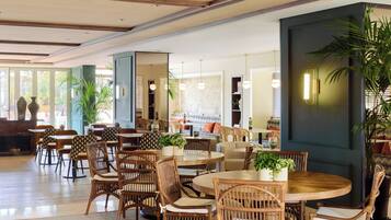 A spacious indoor area with multiple round tables and wicker chairs, surrounded by large windows and indoor plants.