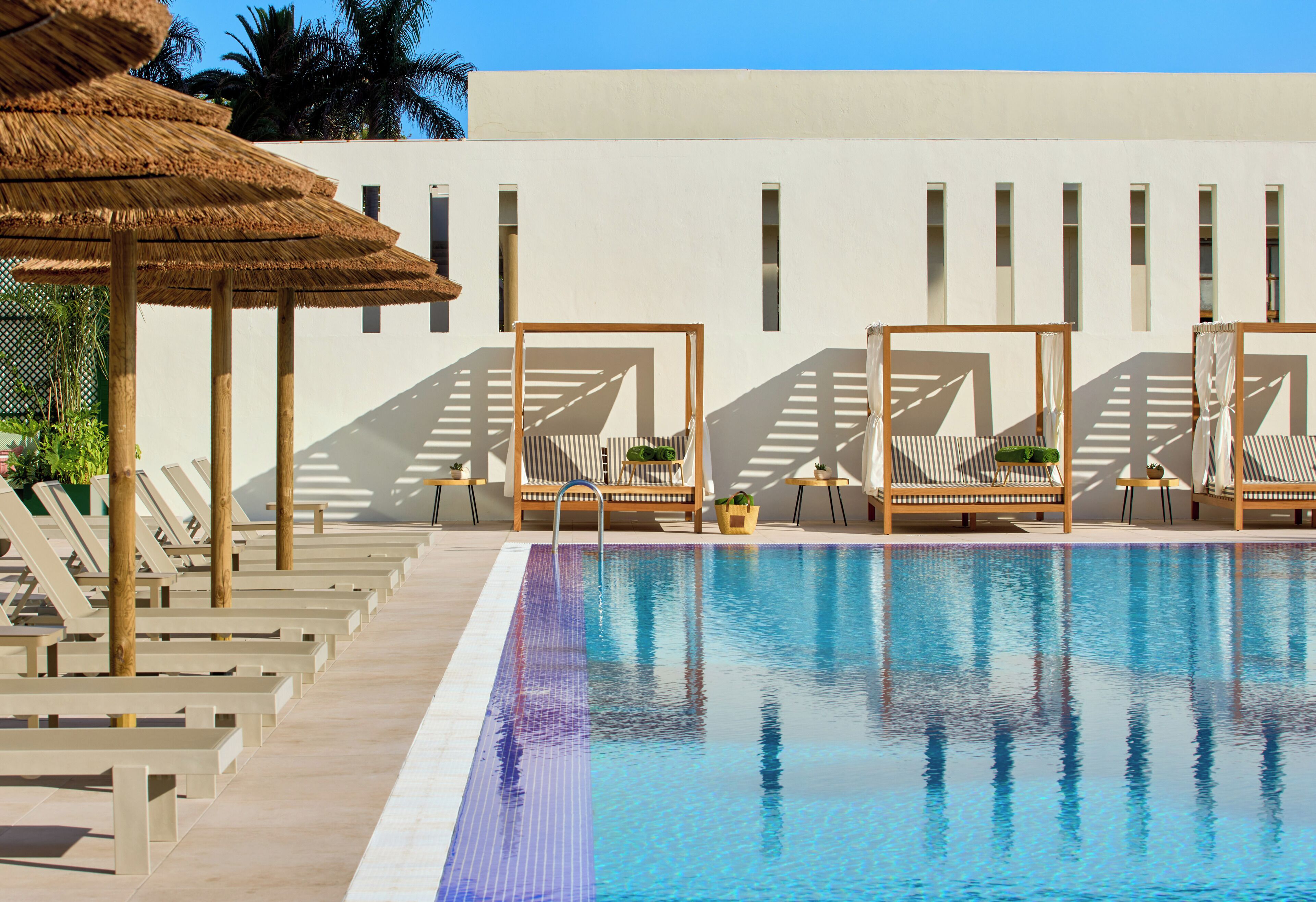 A swimming pool with lounge chairs and umbrellas, surrounded by a white building with windows.