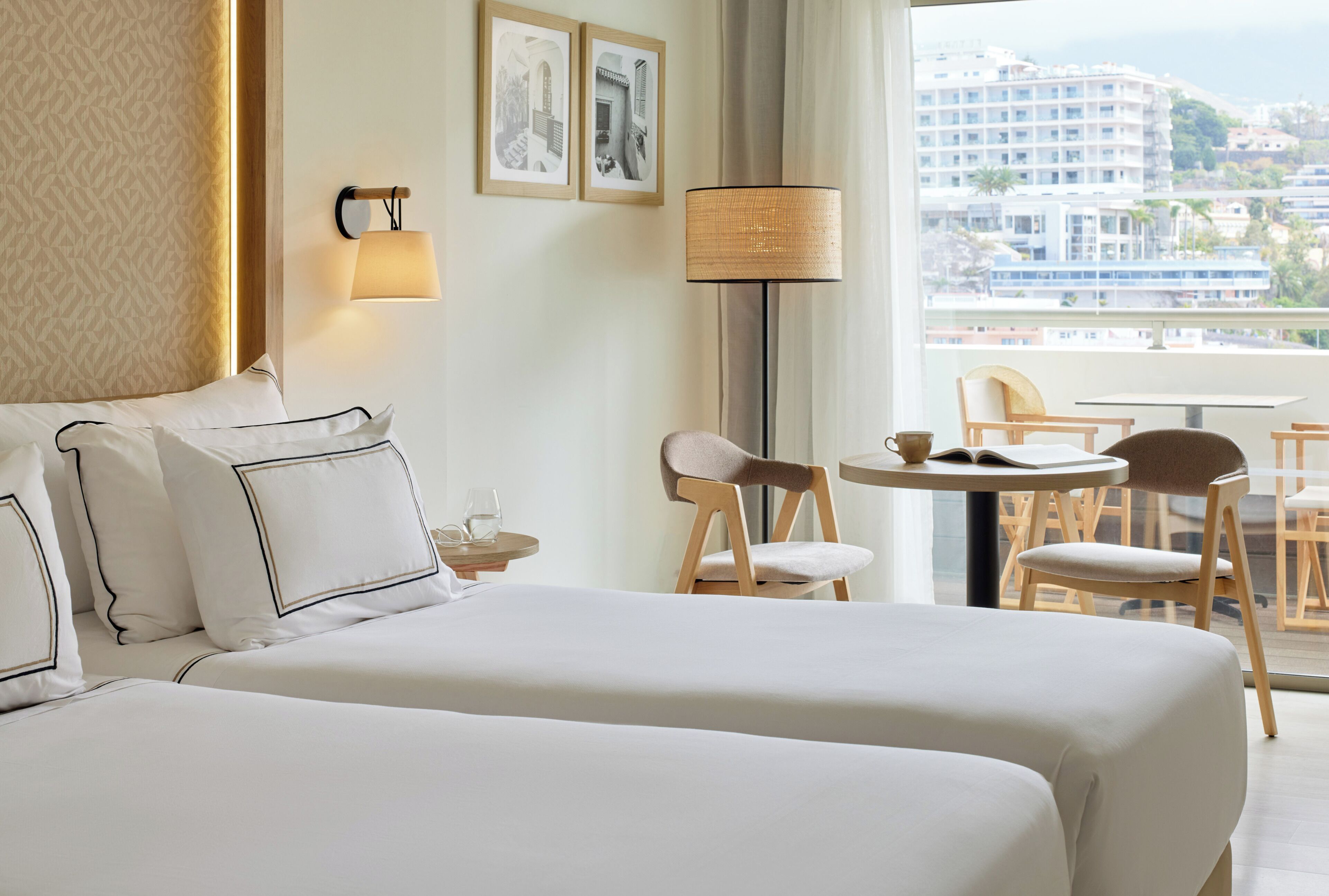 Habitación de hotel con dos camas, una mesita con sillas, una lámpara y vistas a edificios a través de una ventana.