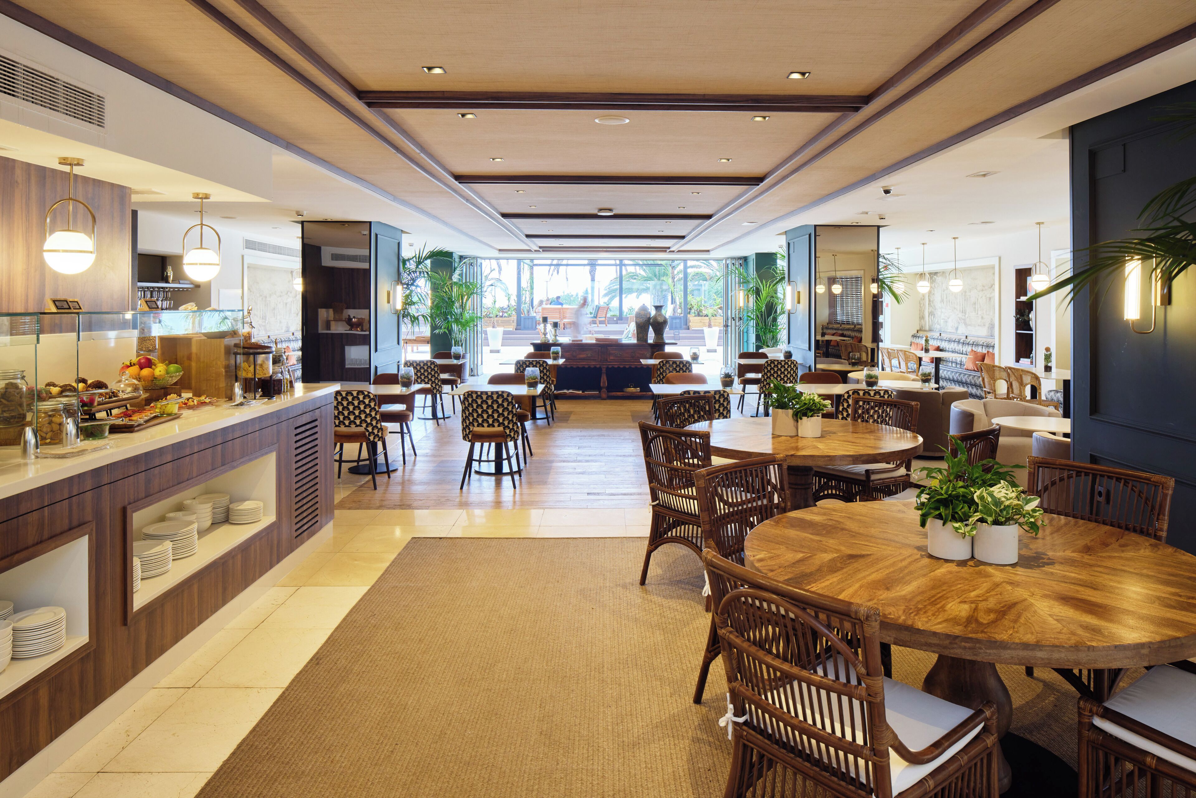 Een ruime eetruimte met een ronde centrale tafel, omringd door rieten stoelen, een buffet met diverse gerechten en een uitzicht op de omgeving door grote ramen.