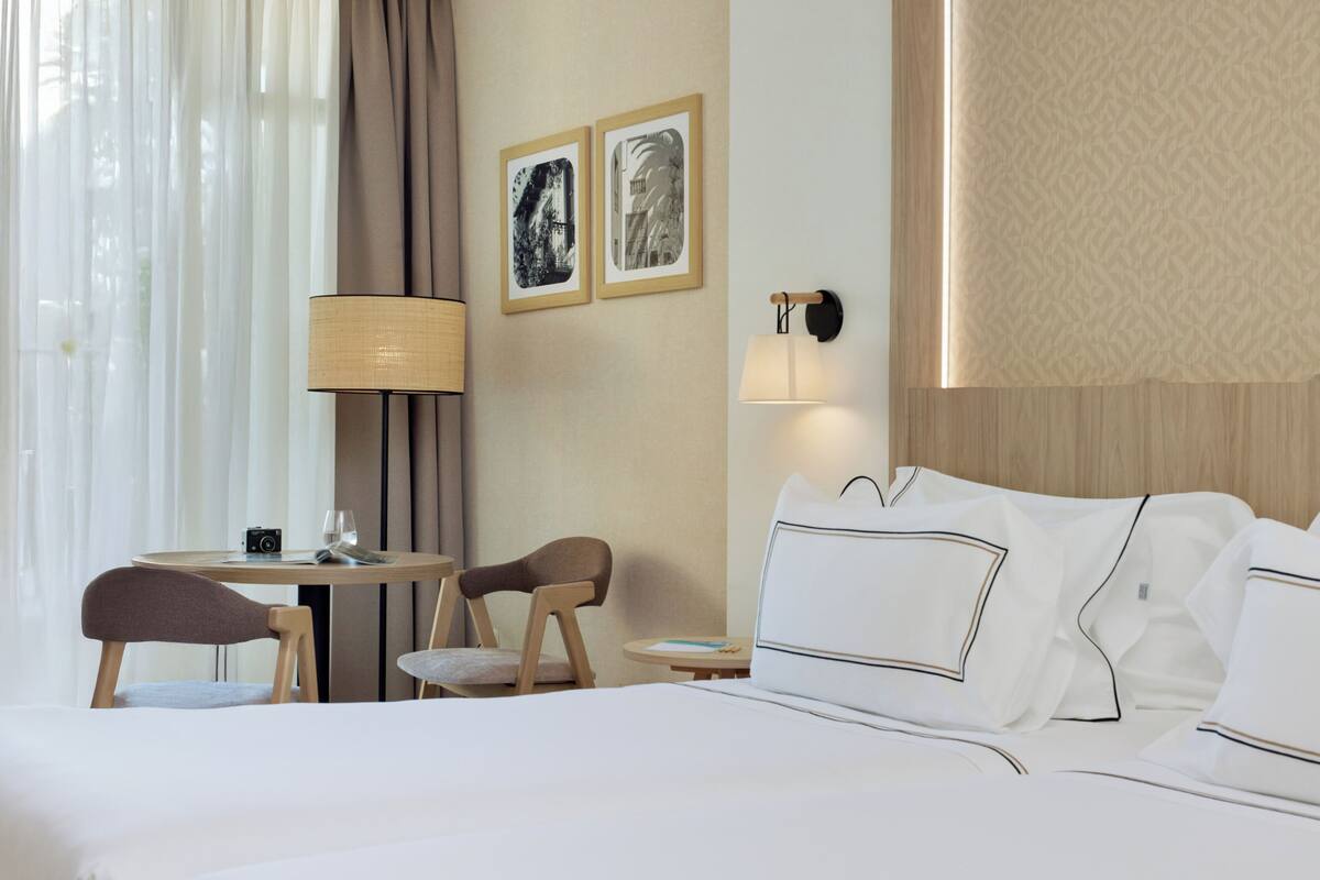 Hotellihuoneessa on sänky, kaksi tuolia, pieni pöytä, lamppu ja kaksi kehystettyä kuvaa seinällä.