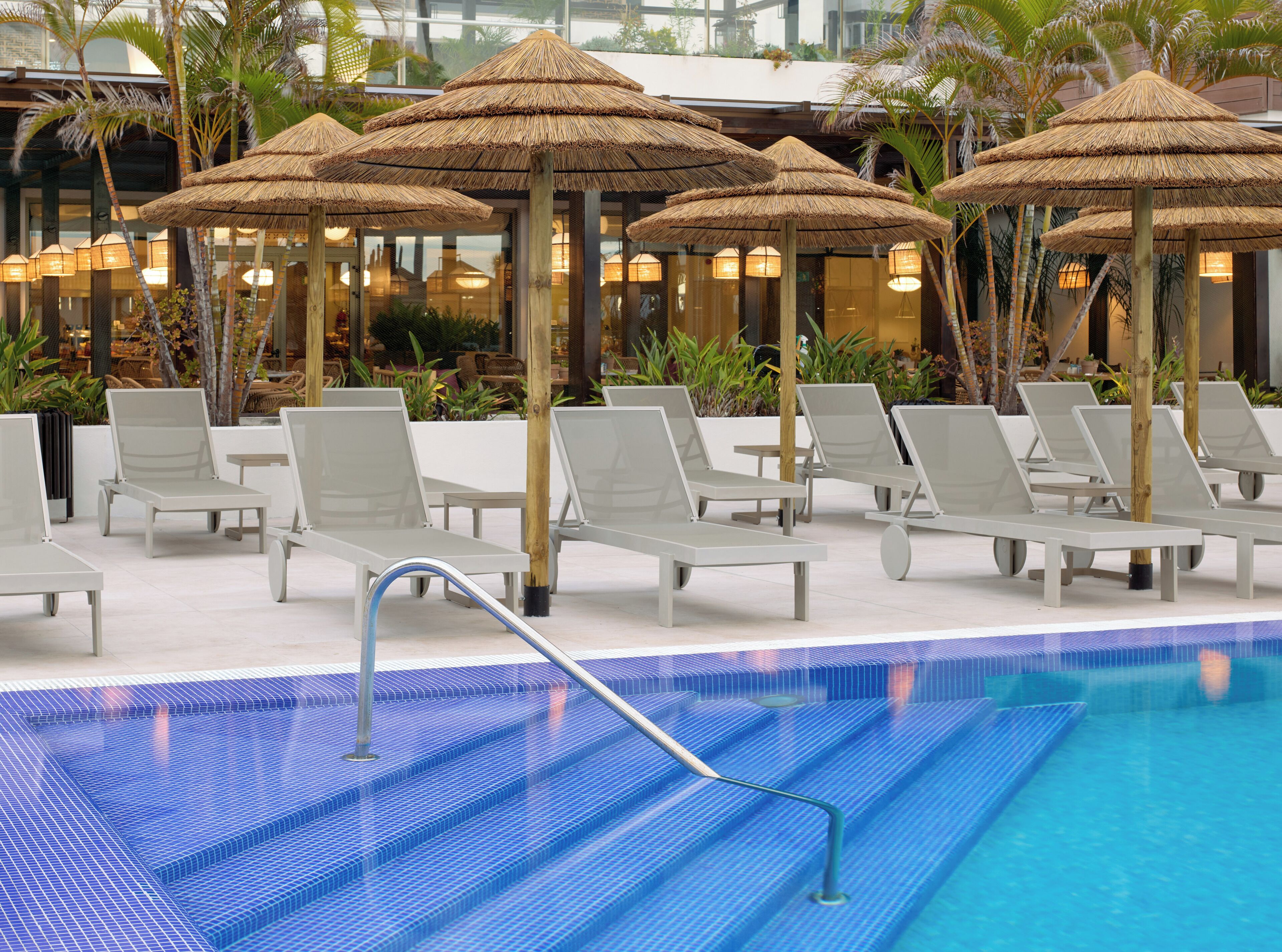 Zona de piscina con sillones de descanso y sombrillas de paja, rodeada de plantas y un edificio al fondo.