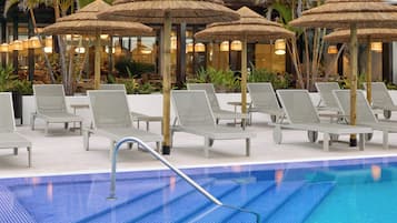 Zona de piscina con sillones de descanso y sombrillas de paja, rodeada de plantas y un edificio al fondo.