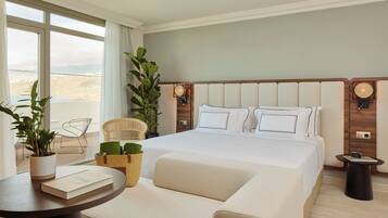 A modern hotel room with a large bed, a round table with a book and a plant, a chair, and a view of the desert through the window.