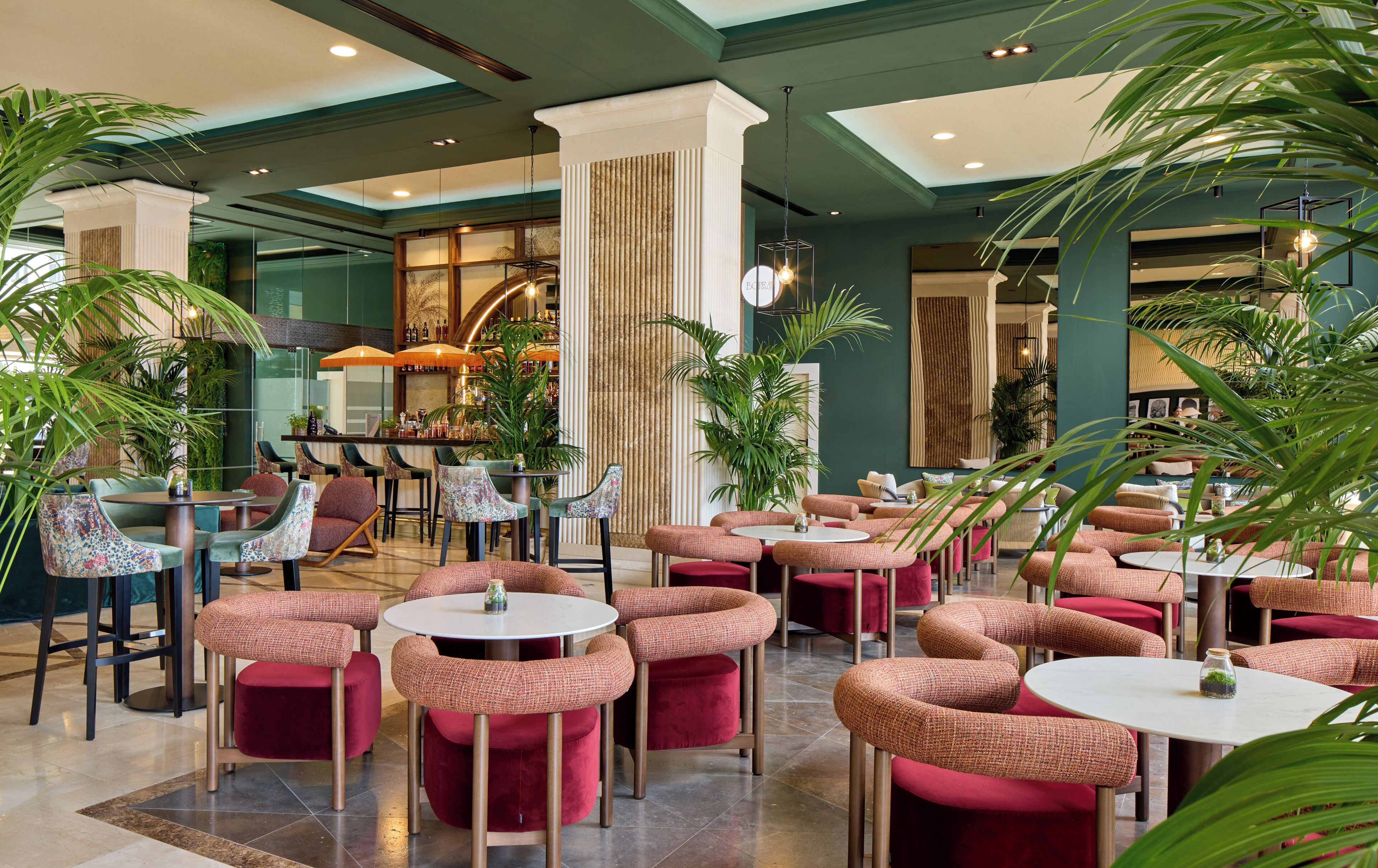 A spacious indoor area with multiple seating arrangements, including round tables and cushioned chairs, surrounded by potted plants.