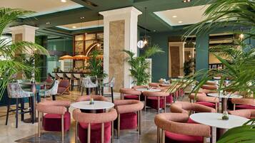 A spacious indoor area with multiple seating arrangements, including round tables and cushioned chairs, surrounded by potted plants.