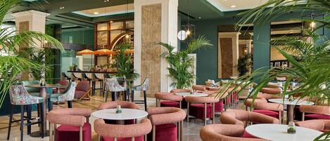 A spacious indoor area with multiple seating arrangements, including round tables and cushioned chairs, surrounded by potted plants.
