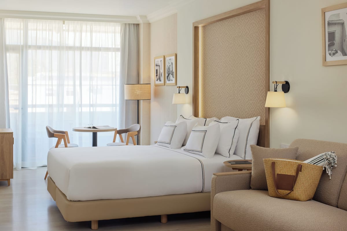 Hotellihuoneessa on suuri sänky, sohva, pieni pöytä ja kaksi tuolia. Seinälle on kiinnitetty kaksi lamppua, ja huoneessa on suuri ikkuna, jossa on ohuet verhot.