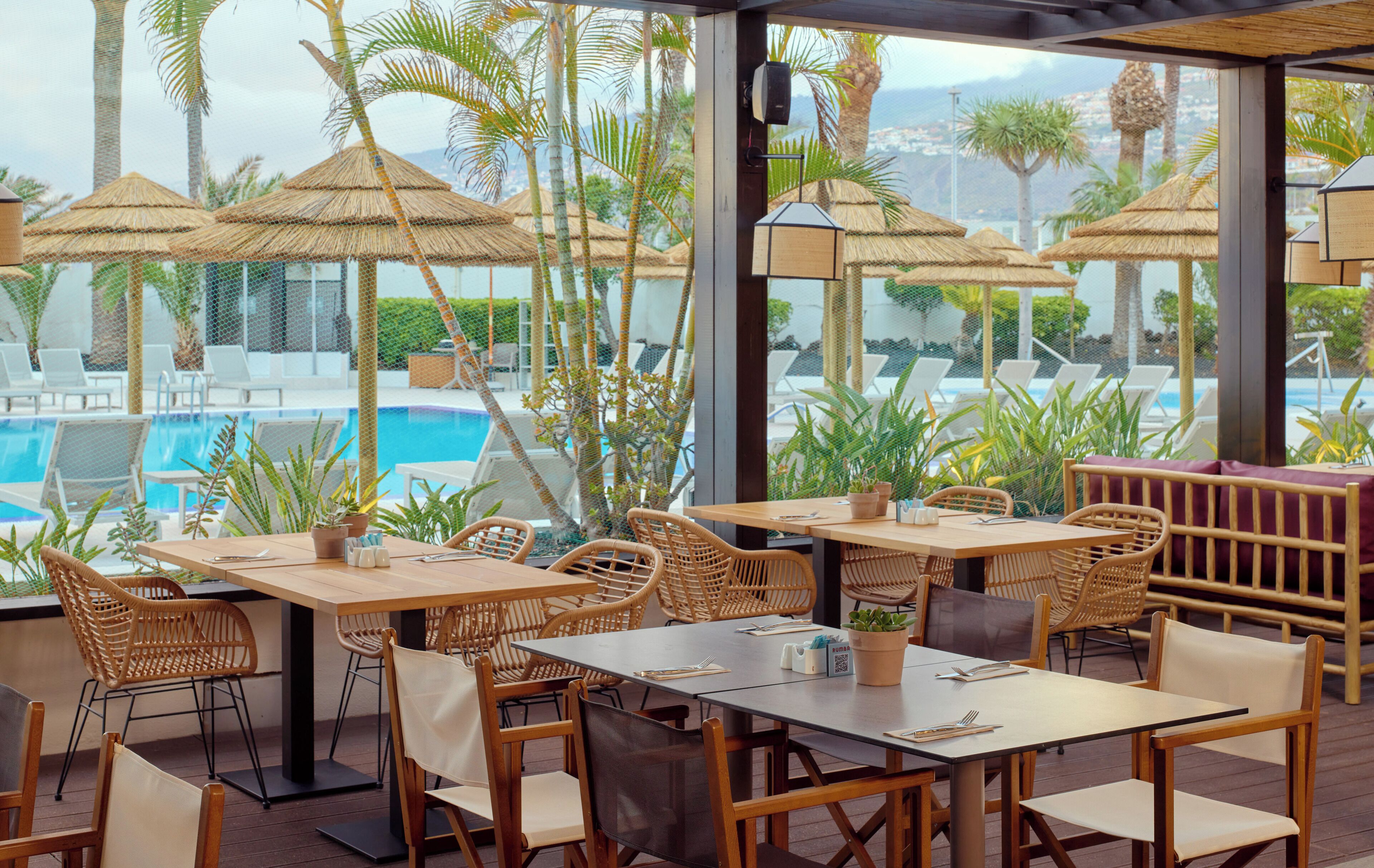 Un área de comedor al aire libre con mesas y sillas, rodeada de sombrillas y plantas en macetas, con vista a una piscina.