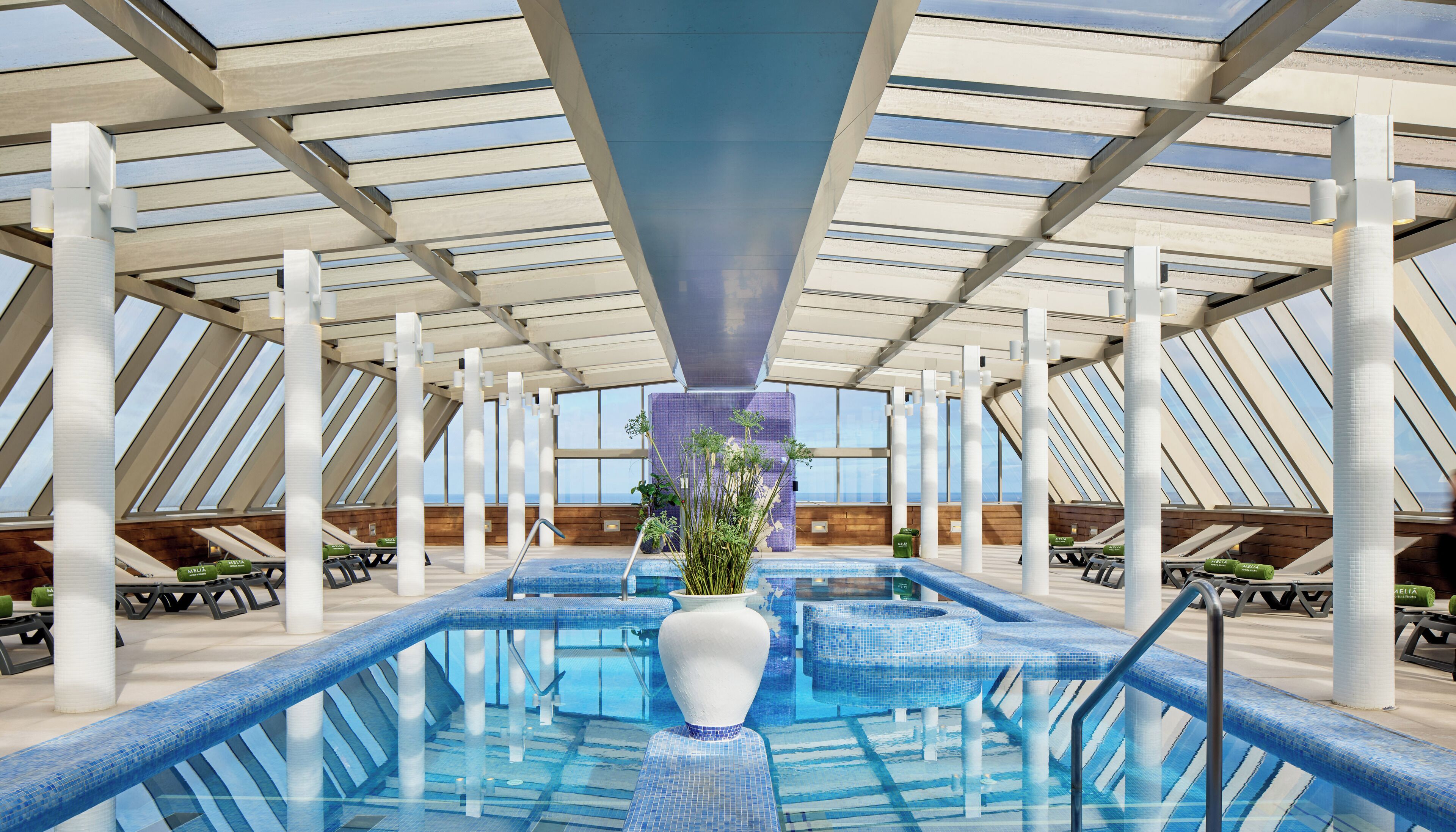 Una piscina interior con diseño moderno, que incluye un jardín vertical central, varias zonas de estar y un techo alto con tragaluces.
