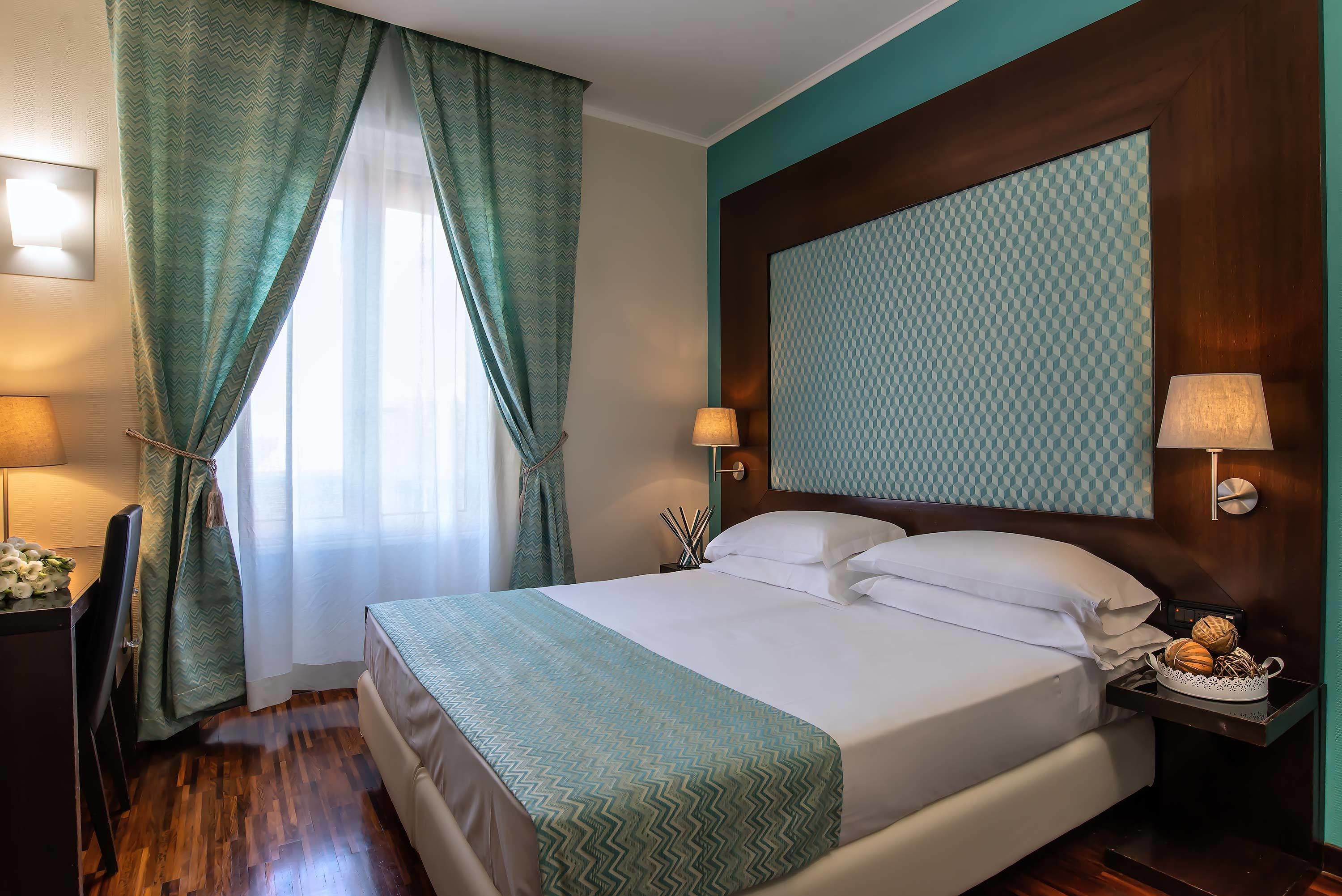 A hotel room with a bed, a desk, and a chair. There are two lamps on the nightstand and a window with curtains.