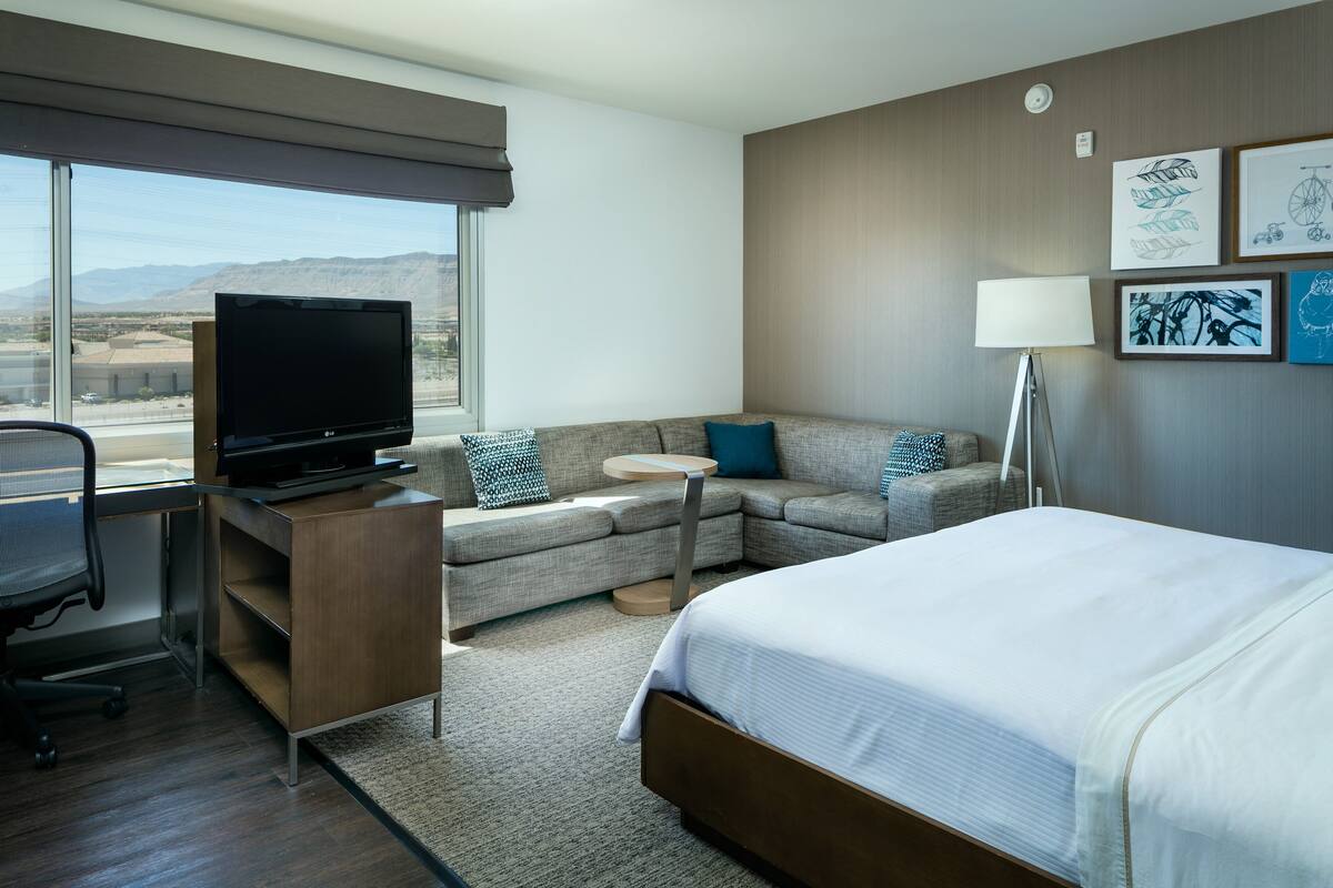 A hotel room with a bed, a sofa, a desk with a TV, a lamp, and framed artwork on the wall.