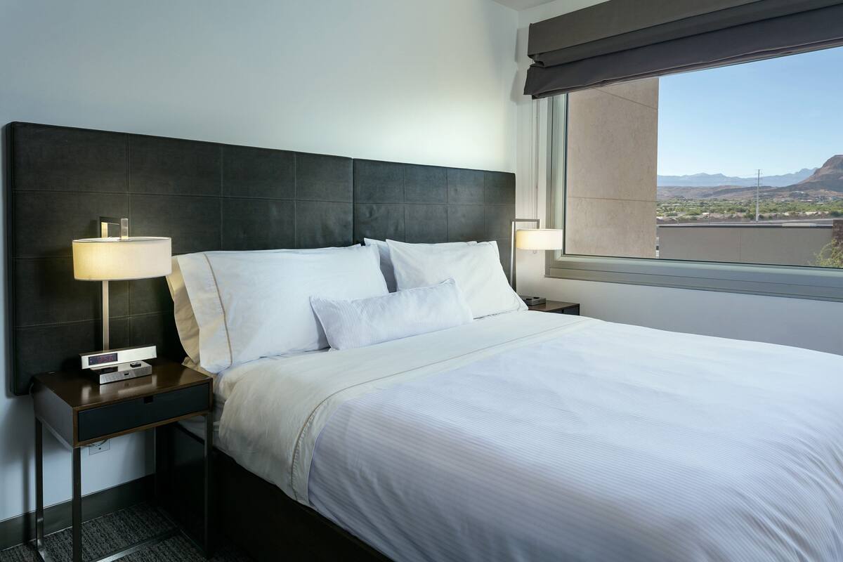 A hotel room with a large bed, two bedside tables, a nightstand, and a window with a view of mountains.