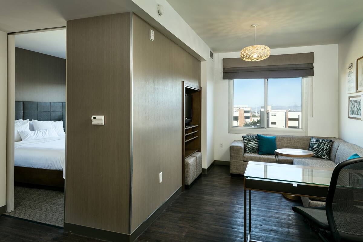A hotel room with a bed, a seating area with a sofa and armchairs, a small table, and a view of buildings through a window.