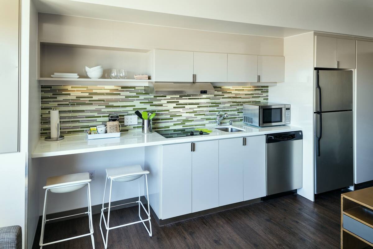A modern kitchen with white cabinets, a stainless steel refrigerator, a microwave, and a dishwasher. There are two white bar stools and a green plant on the counter.