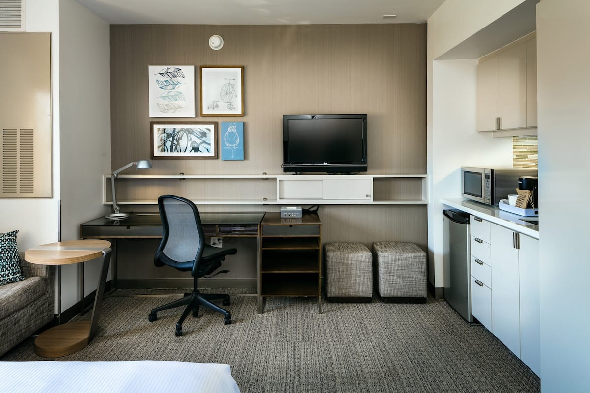 A hotel room with a bed, a desk with a computer, a TV, a small kitchenette, and a sofa.