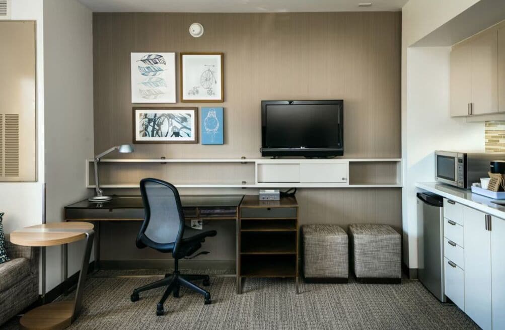 Een kleine hotelkamer met een bureau, stoel, televisie, kitchenette en kunstwerk aan de muur.