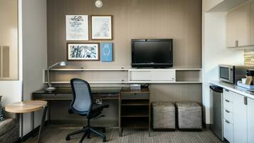 Een kleine hotelkamer met een bureau, stoel, televisie, kitchenette en kunstwerk aan de muur.