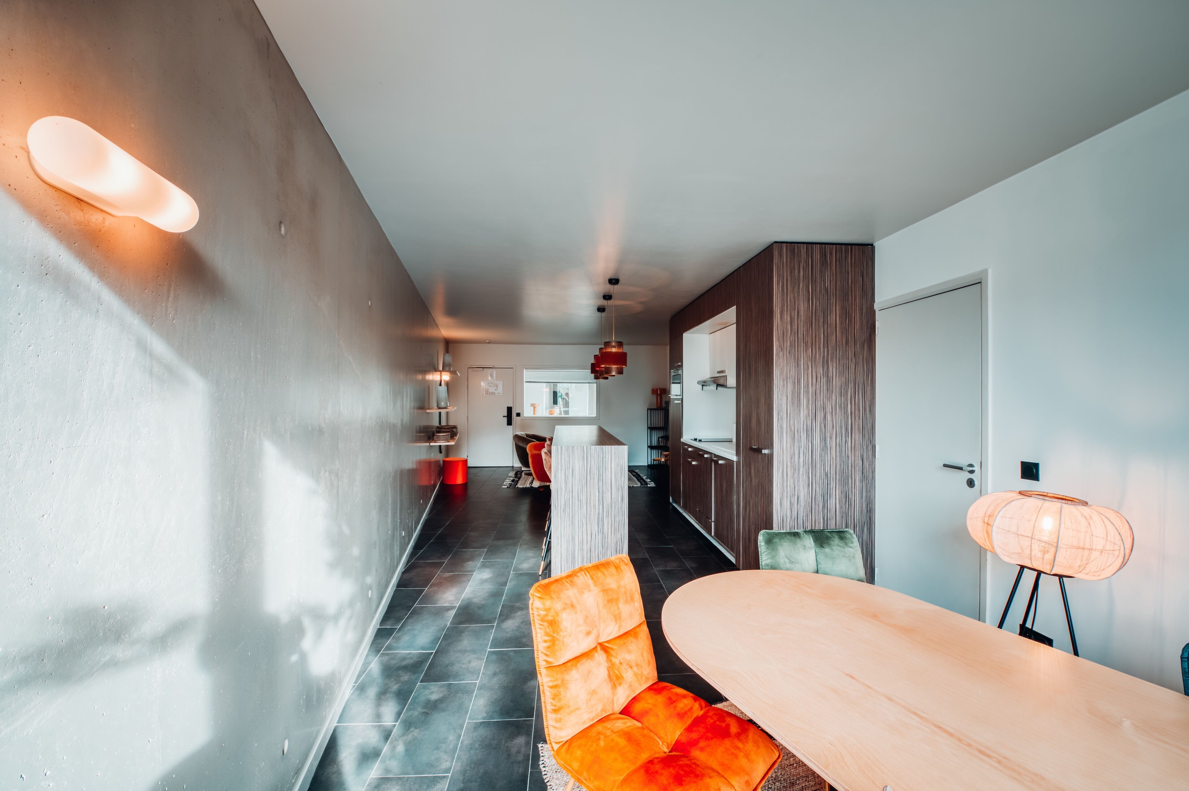 A modern kitchen with dark tiles, a wooden cabinet, and a dining area with a table and orange chairs.