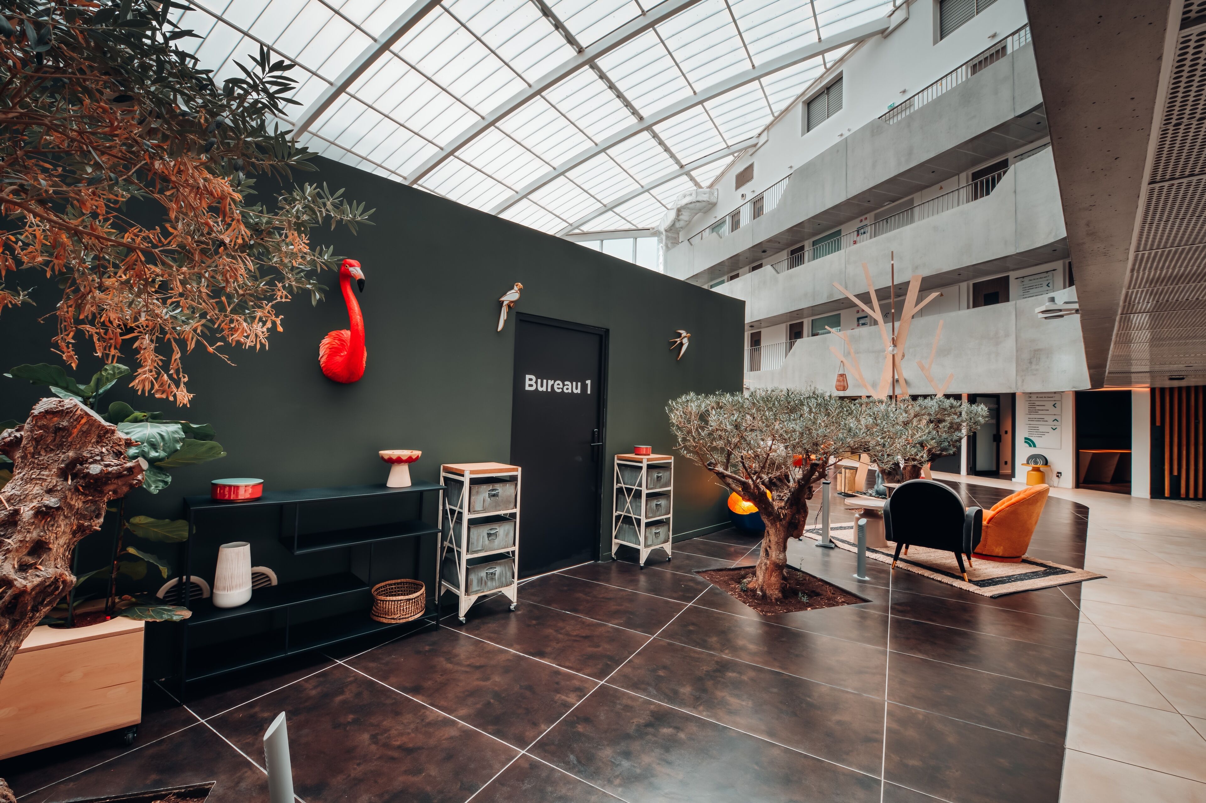 A modern interior with a high ceiling, a sign reading "Bureau 1", and a seating area with a tree and orange chairs.