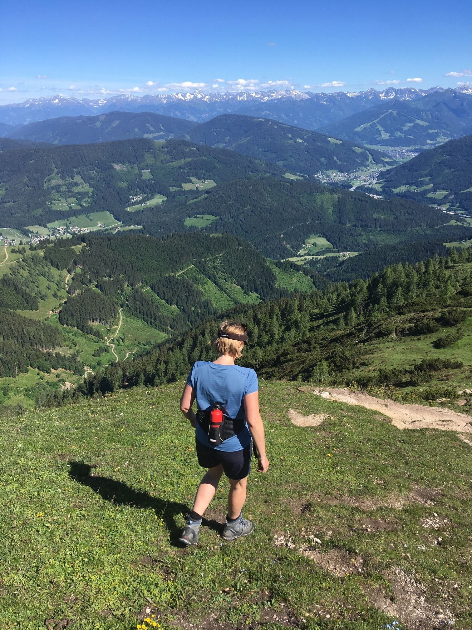Une personne fait de la randonnée sur une colline herbeuse, un sac à dos sur le dos, et contemple une vallée avec des montagnes à l’horizon.
