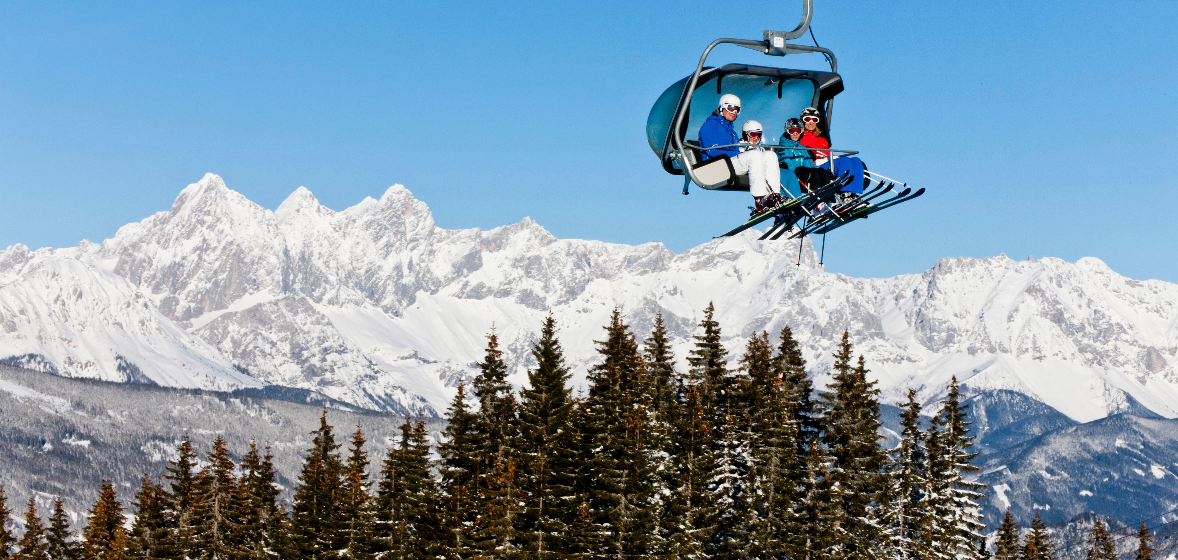 A ski lift with three skiers in blue and red jackets, carrying skis and poles, against a backdrop of snow-covered mountains and coniferous trees.