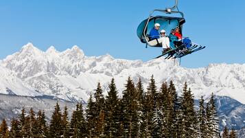 A ski lift with three skiers in blue and red jackets, carrying skis and poles, against a backdrop of snow-covered mountains and coniferous trees.