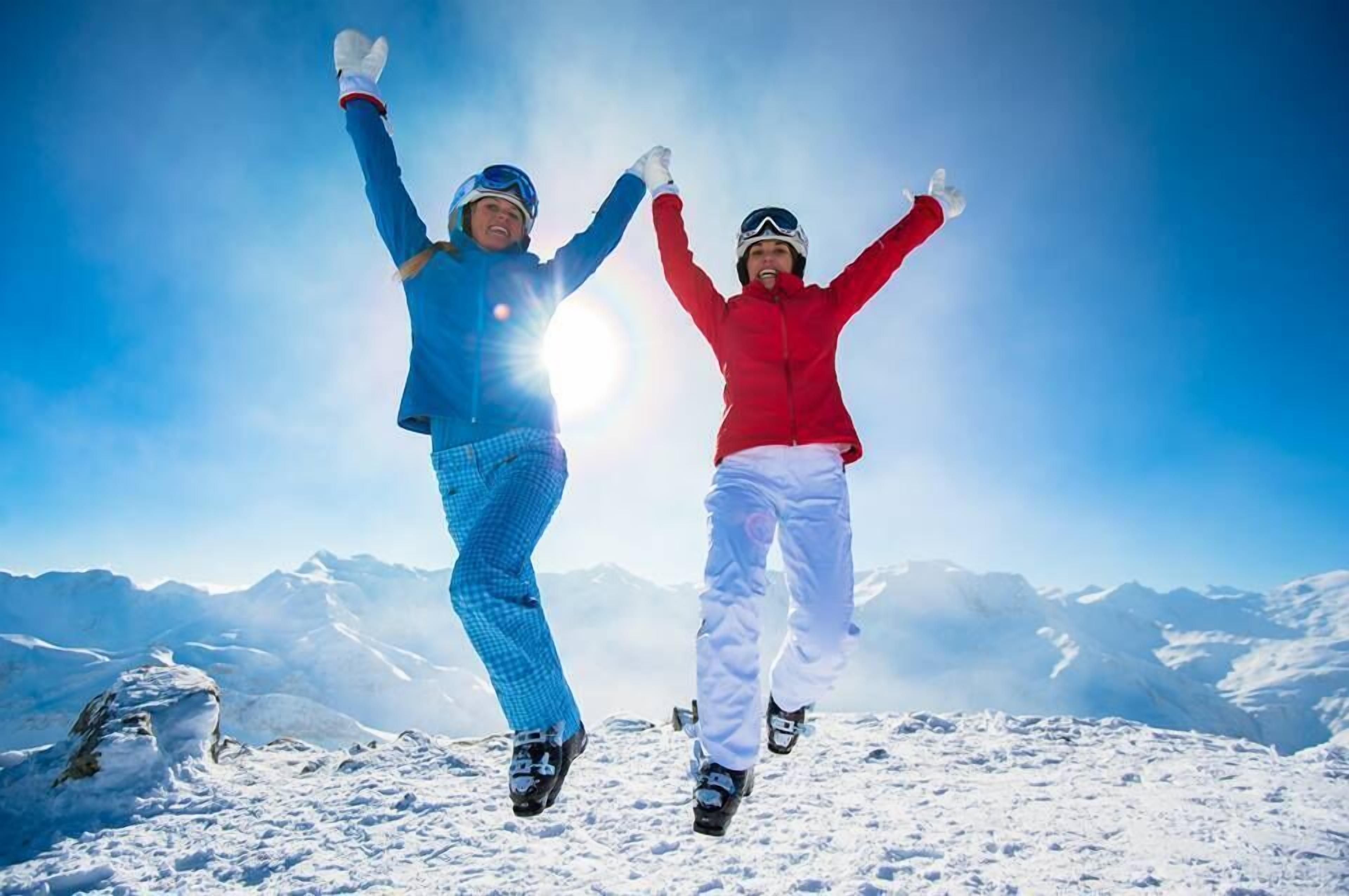 Dos personas saltando en una montaña nevada bajo un cielo despejado, con montañas lejanas al fondo.