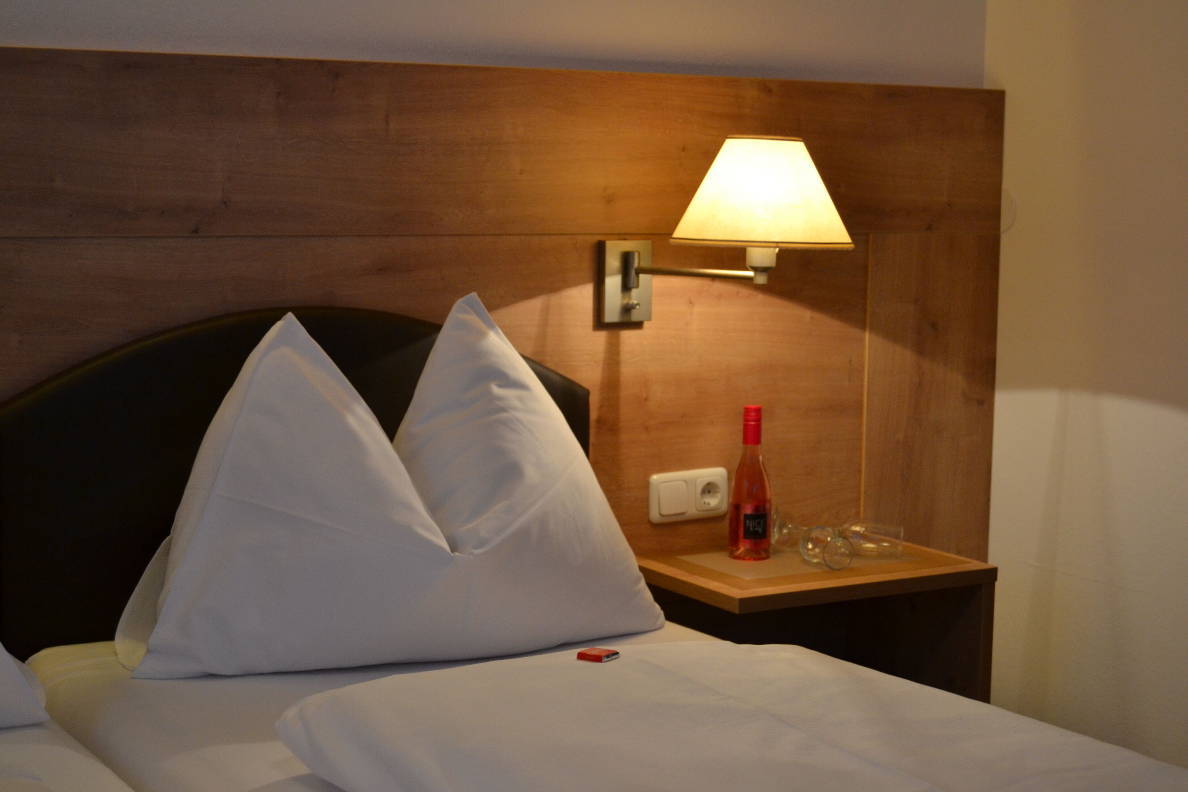 Une chambre d’hôtel avec une tête de lit en bois, deux oreillers blancs, une table de chevet avec une lampe, une bouteille et un verre.
