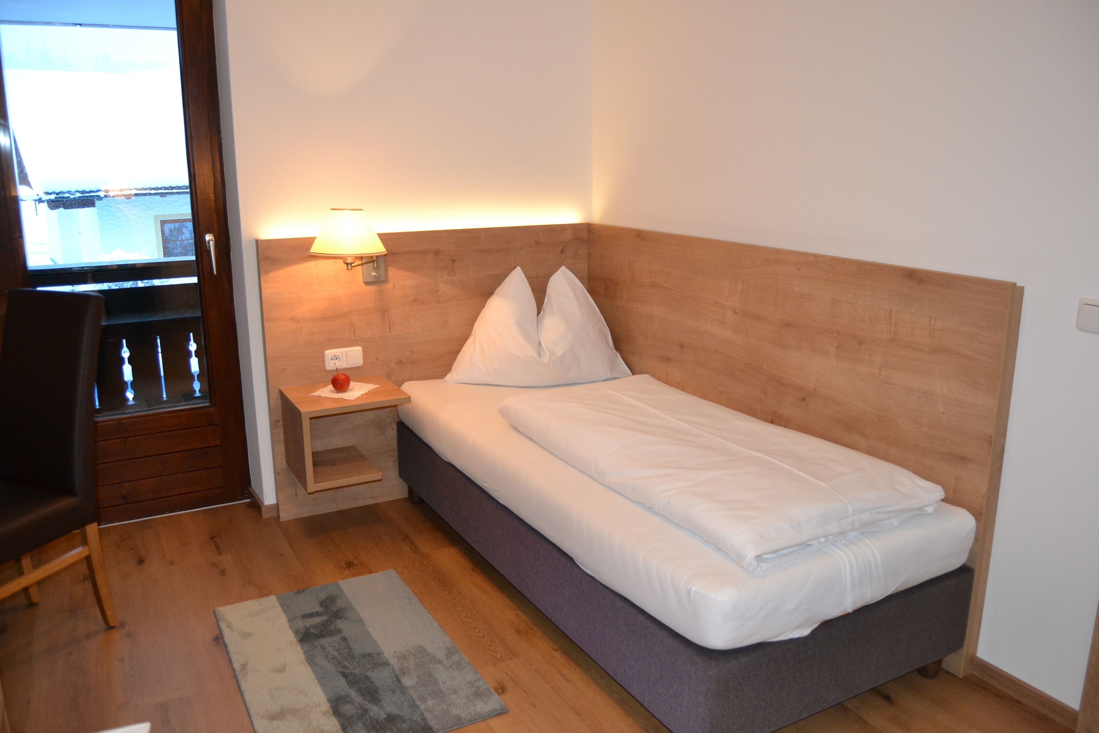 Ein kleines, ordentlich bezogenes Bett mit einem hölzernen Kopfteil, ein Nachttisch mit einer Lampe und Blick auf einen Balkon durch ein Fenster.
