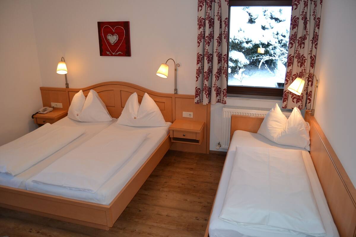 Hotelkamer met twee bedden, nachtlampjes, een nachtkastje met een telefoon, een radiator en een raam met gordijnen.