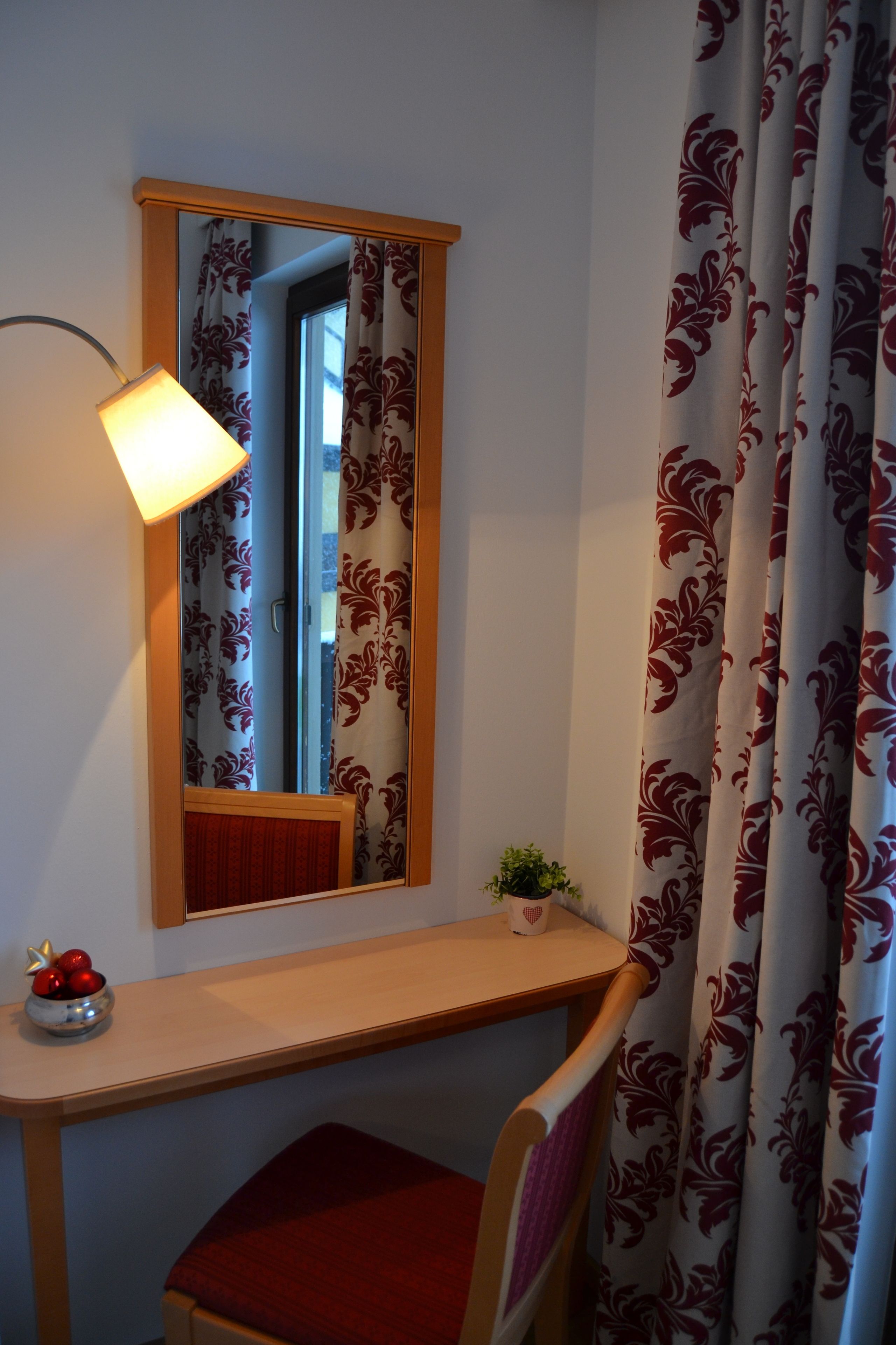 A room with a wooden desk, a chair, a small table with a plant, a mirror, and a window with curtains.
