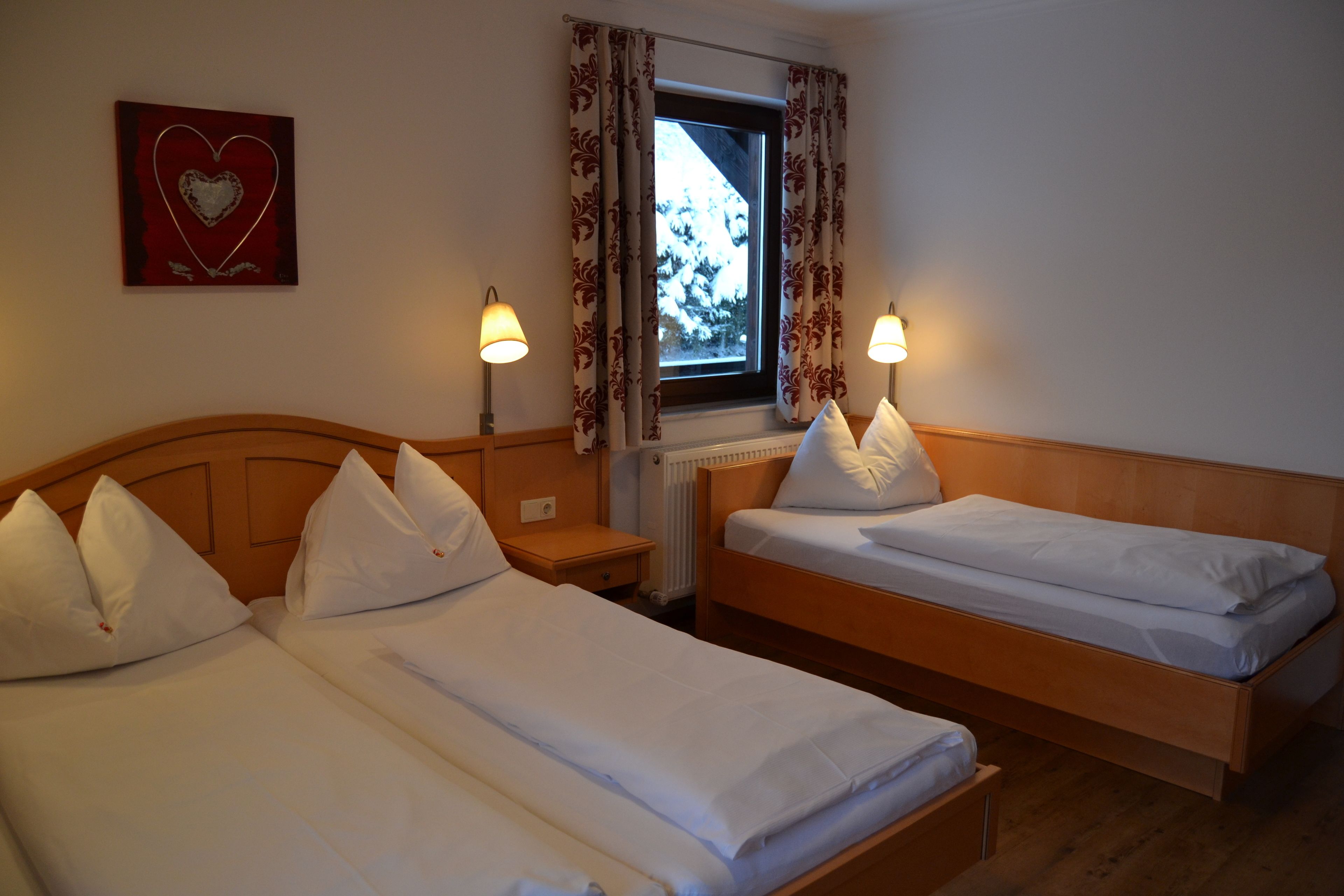 Een hotelkamer met twee enkele bedden, een raam met gordijnen, een hartdecoratie aan de muur en twee bedlampjes.
