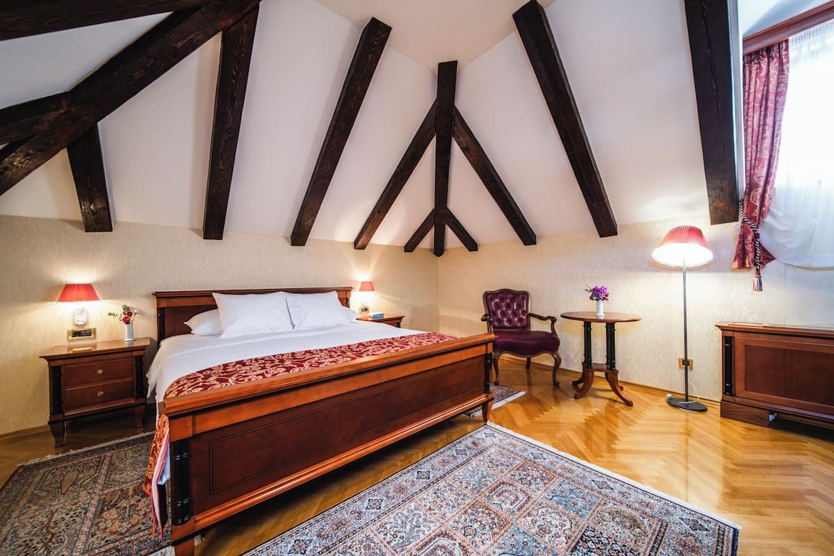 Ein Schlafzimmer mit einem großen Holzbett, zwei Nachttischen mit Lampen, einem Sessel, einem kleinen runden Tisch mit einer Vase und einem großen Fenster mit Vorhängen.